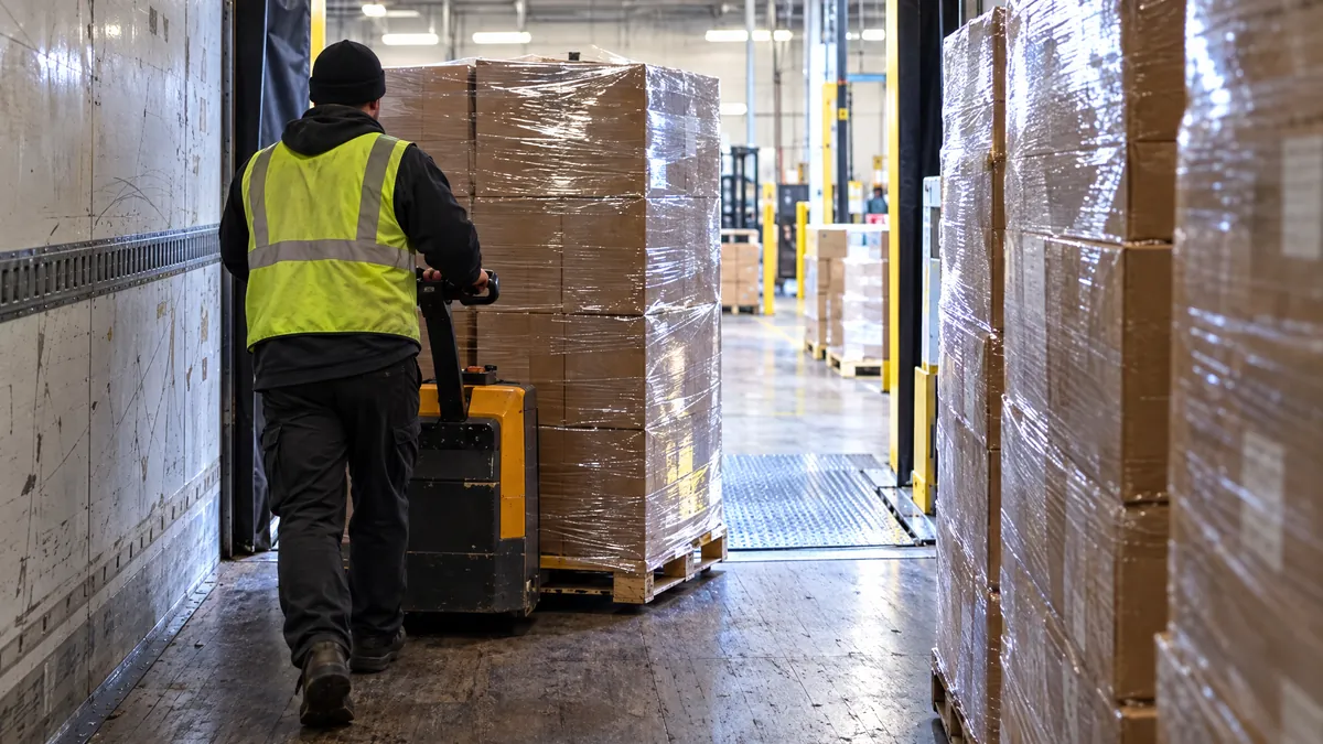 View from inside a trailer: a worker moves a stretch-wrapped pallet of cartons with a pallet jack